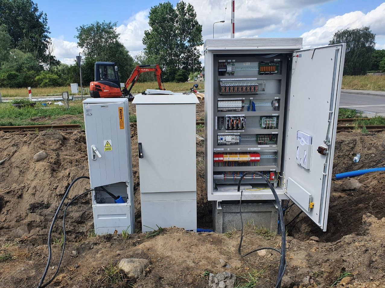 Otwarta szafa sterownicza z widocznymi elementami instalacji elektrycznej, okablowaniem i zabezpieczeniami, umieszczona na zewnątrz w ziemi, z małą koparką w tle, sugerująca prace instalacyjne w toku.
