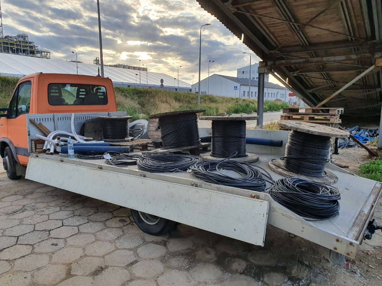 Pomarańczowy samochód dostawczy w Raba Niżna z nawiniętymi kablami na drewnianych szpulach i rurami na platformie, przygotowany do prac instalacyjnych, widok na budynki przemysłowe w tle.