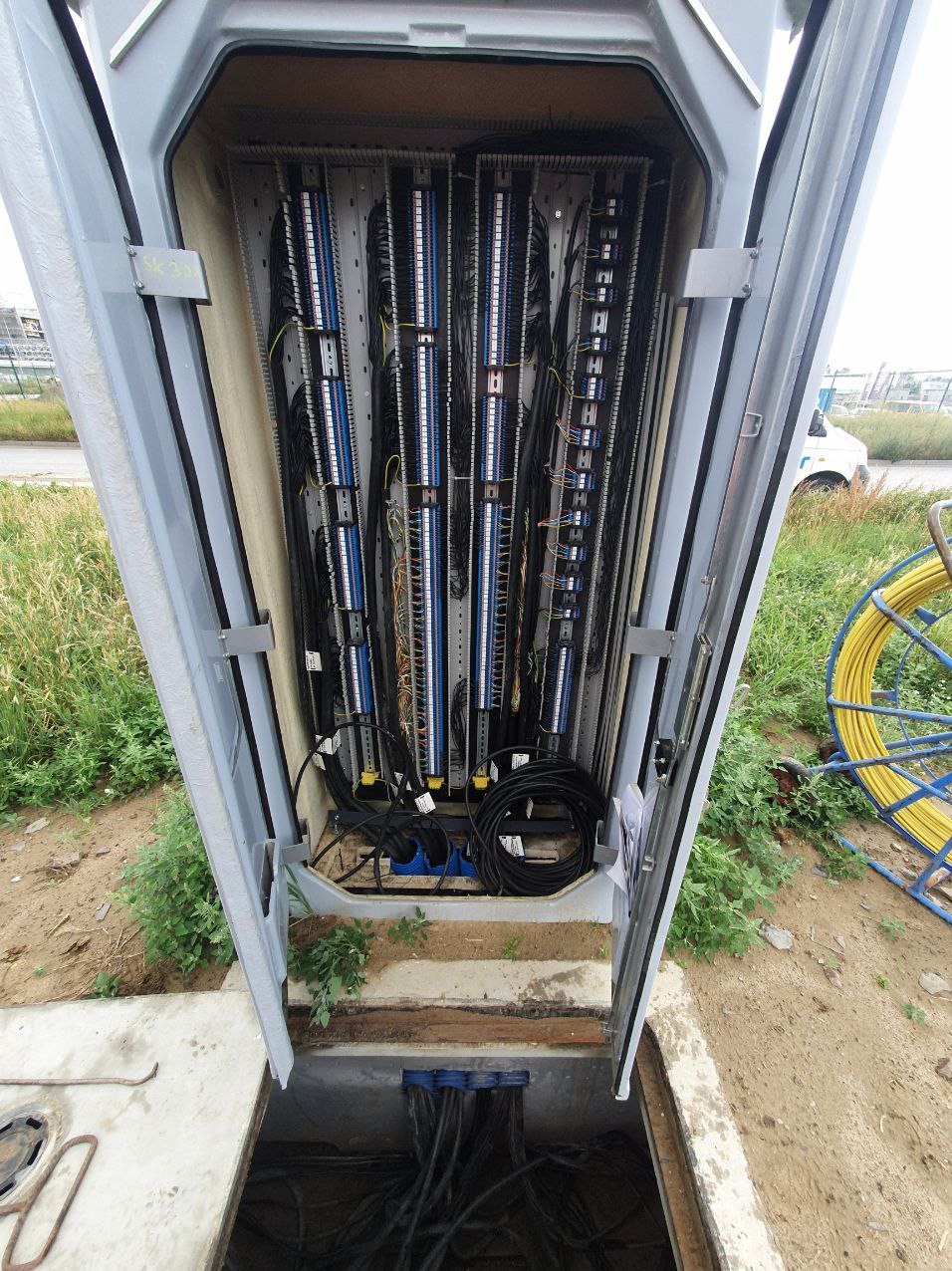 Otwarta skrzynka telekomunikacyjna z widocznymi kablami, złączami i panelami krosowniczymi, z boku widoczny bęben z kablem światłowodowym.