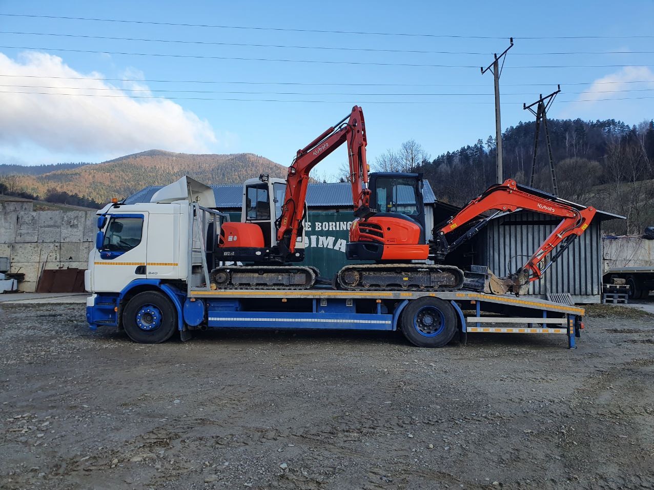 Ciężarówka z dwoma pomarańczowymi minikoparkami Kubota na platformie, w tle widoczne góry i betonowe bloki.