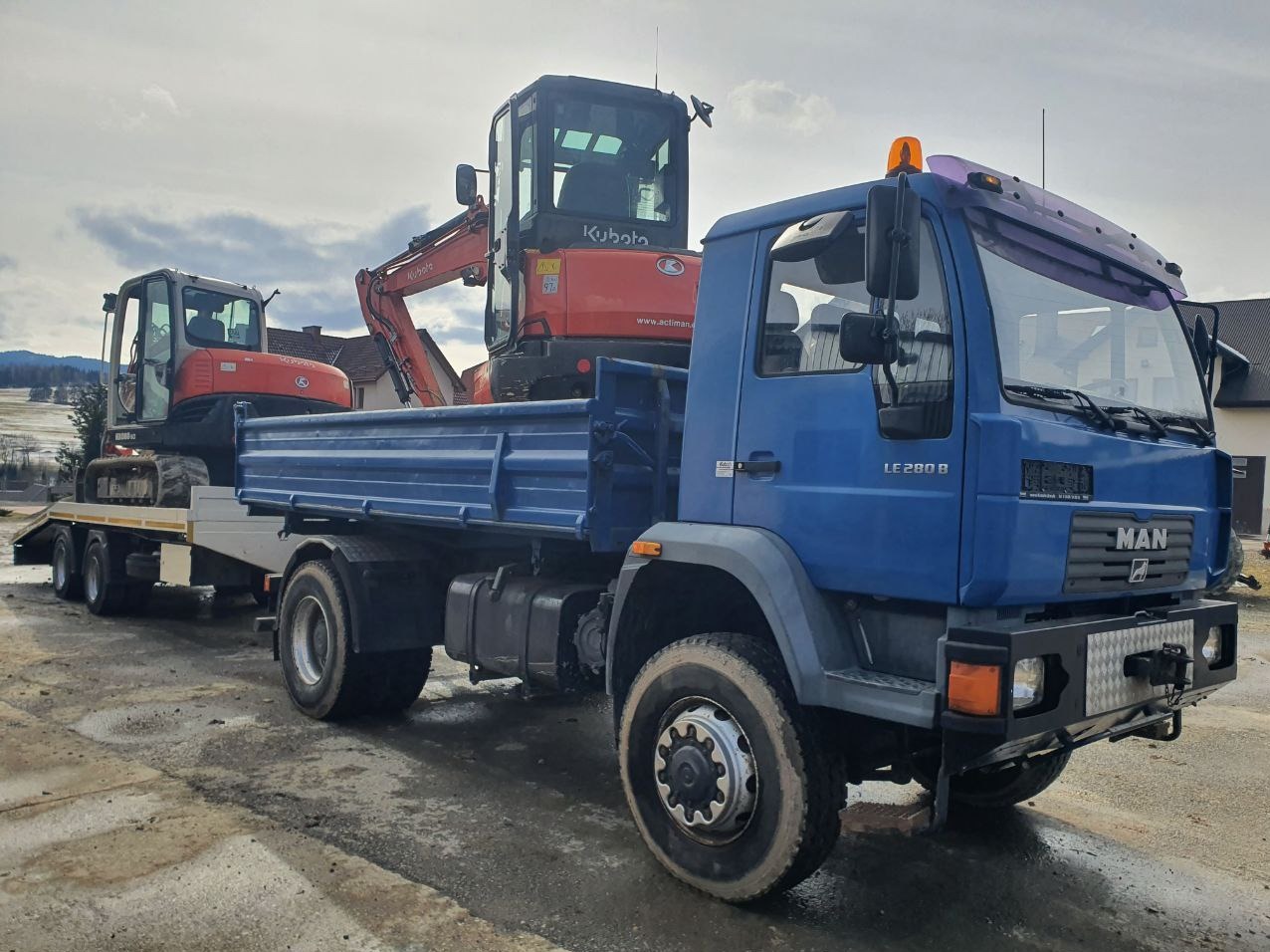Niebieska ciężarówka MAN z przyczepą, transportująca dwie pomarańczowe mini koparki Kubota na placu budowy, widoczne góry w tle.