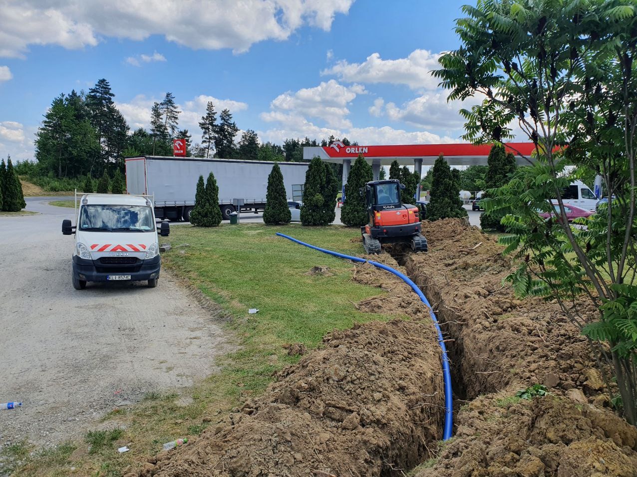 Pomarańczowa mini koparka Kubota przy wykopie z niebieską rurą, na tle stacji Orlen i białego samochodu dostawczego.