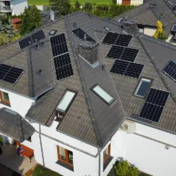 Widok z góry na dom jednorodzinny z zainstalowanymi panelami słonecznymi na dachu z dachówki ceramicznej, widoczne okna dachowe i komin z cegły klinkierowej.