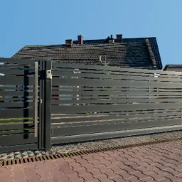 Nowoczesna, przesuwna brama wjazdowa z poziomych profili metalowych, z widocznym domofonem i uchwytem, na tle brukowanej posesji i domu z ceglanym dachem.