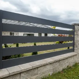 Nowoczesne ogrodzenie z poziomych, szarych paneli zamocowanych między słupkami z szarego kamienia, na niskiej podmurówce z szarej kostki, z widokiem na dom w tle.
