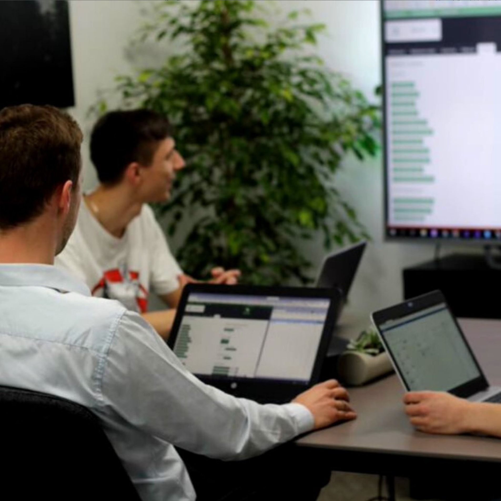 Zespół w trakcie warsztatów, pracujący przy laptopach z widocznymi danymi na ekranach, duży monitor z wykresami w tle, roślina doniczkowa.