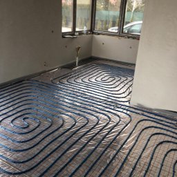 INSTALEKSPERT Kacper Korczykowski - Rozłożone niebieskie rurki ogrzewania podłogowego na folii z czerwoną siatką w surowym pomieszczeniu z oknami.