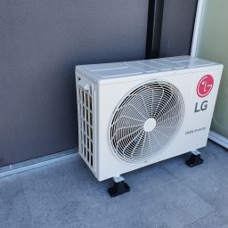 INSTALEKSPERT Kacper Korczykowski - Zewnętrzna jednostka klimatyzatora LG Dual Inverter zamontowana na czarnych podstawkach na szarym, płytkowym balkonie, widok z boku.