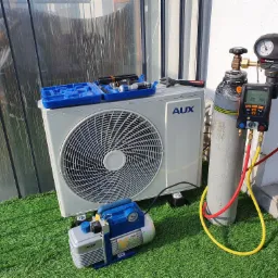 Biała jednostka zewnętrzna klimatyzacji AUX z podłączonymi manometrami i pompą próżniową VALUE na sztucznej trawie, obok butla z gazem i otwarty niebieski organizer z narzędziami.