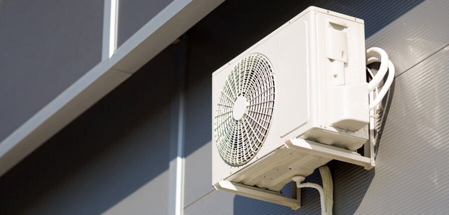 Biała jednostka zewnętrzna klimatyzatora zamontowana na szarej ścianie budynku z widocznymi rurami odprowadzającymi skropliny i przewodami chłodniczymi.