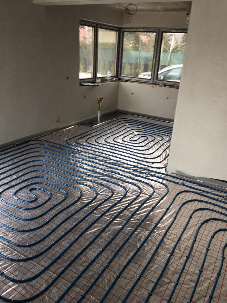 Rozłożone niebieskie rurki ogrzewania podłogowego na folii z czerwoną siatką w surowym pomieszczeniu z oknami.