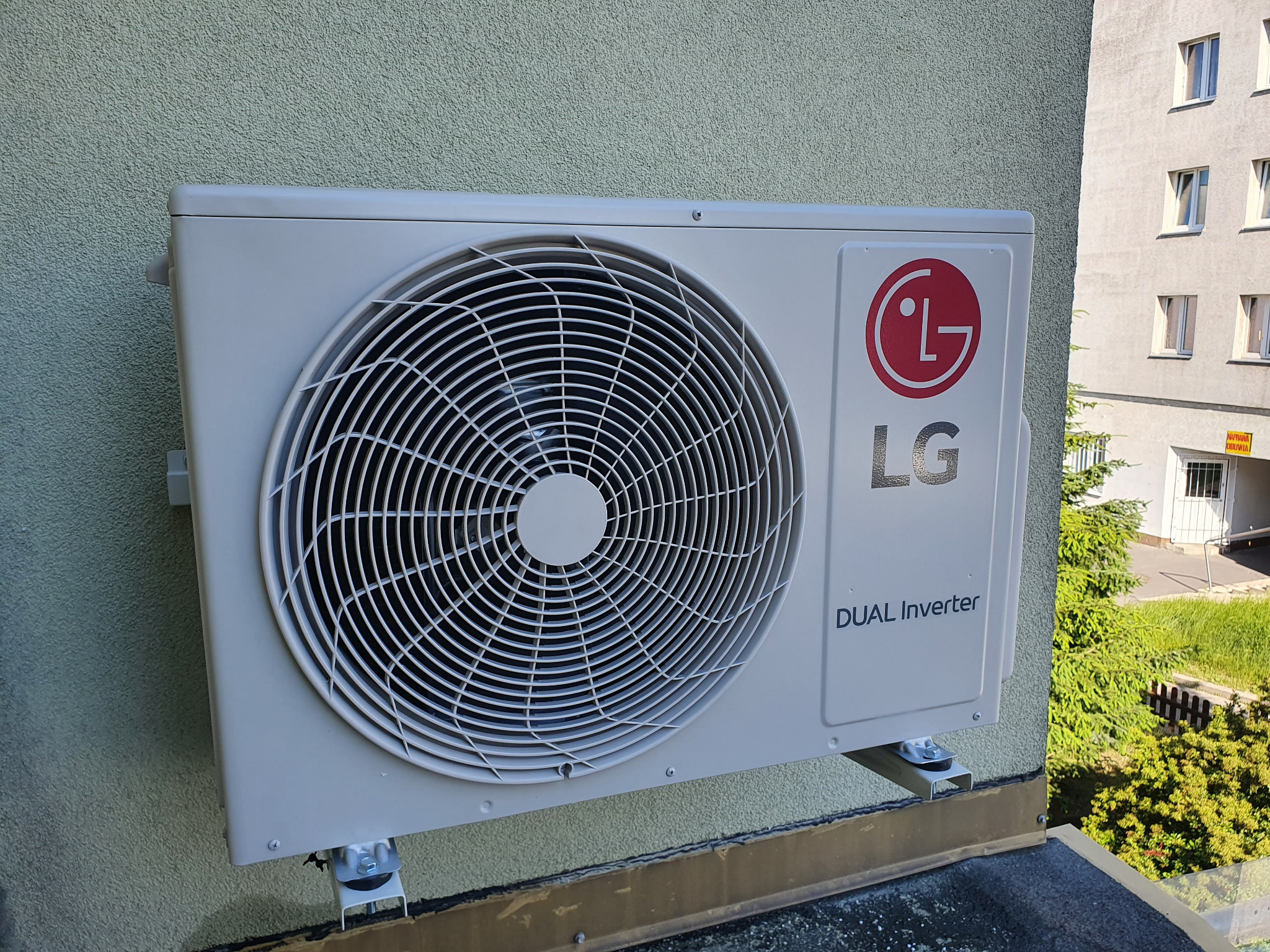 Zewnętrzna jednostka klimatyzacji LG Dual Inverter zamontowana na elewacji budynku, widoczne mocowania i fragment dachu, w tle budynek wielorodzinny i zieleń.