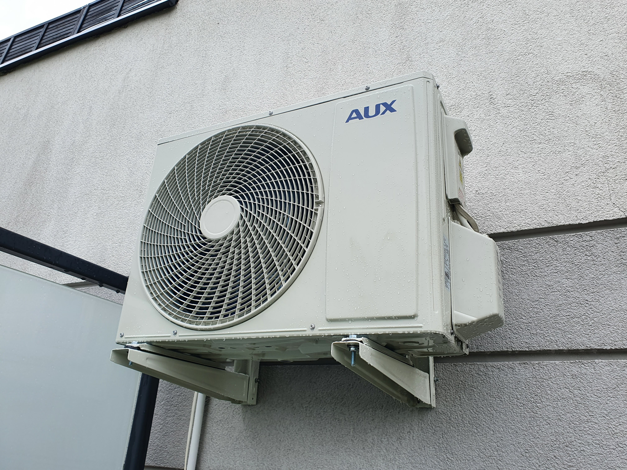 Zewnętrzna jednostka klimatyzatora AUX zamontowana na szarej, teksturowanej ścianie budynku, widoczne krople wody po deszczu.