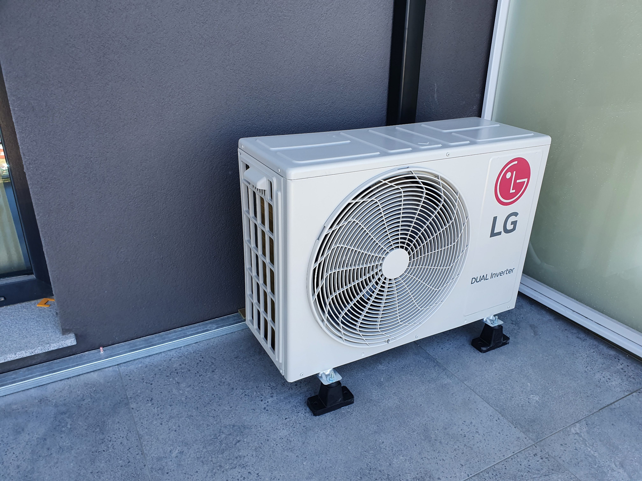 Zewnętrzna jednostka klimatyzatora LG Dual Inverter zamontowana na czarnych podstawkach na szarym, płytkowym balkonie, widok z boku.