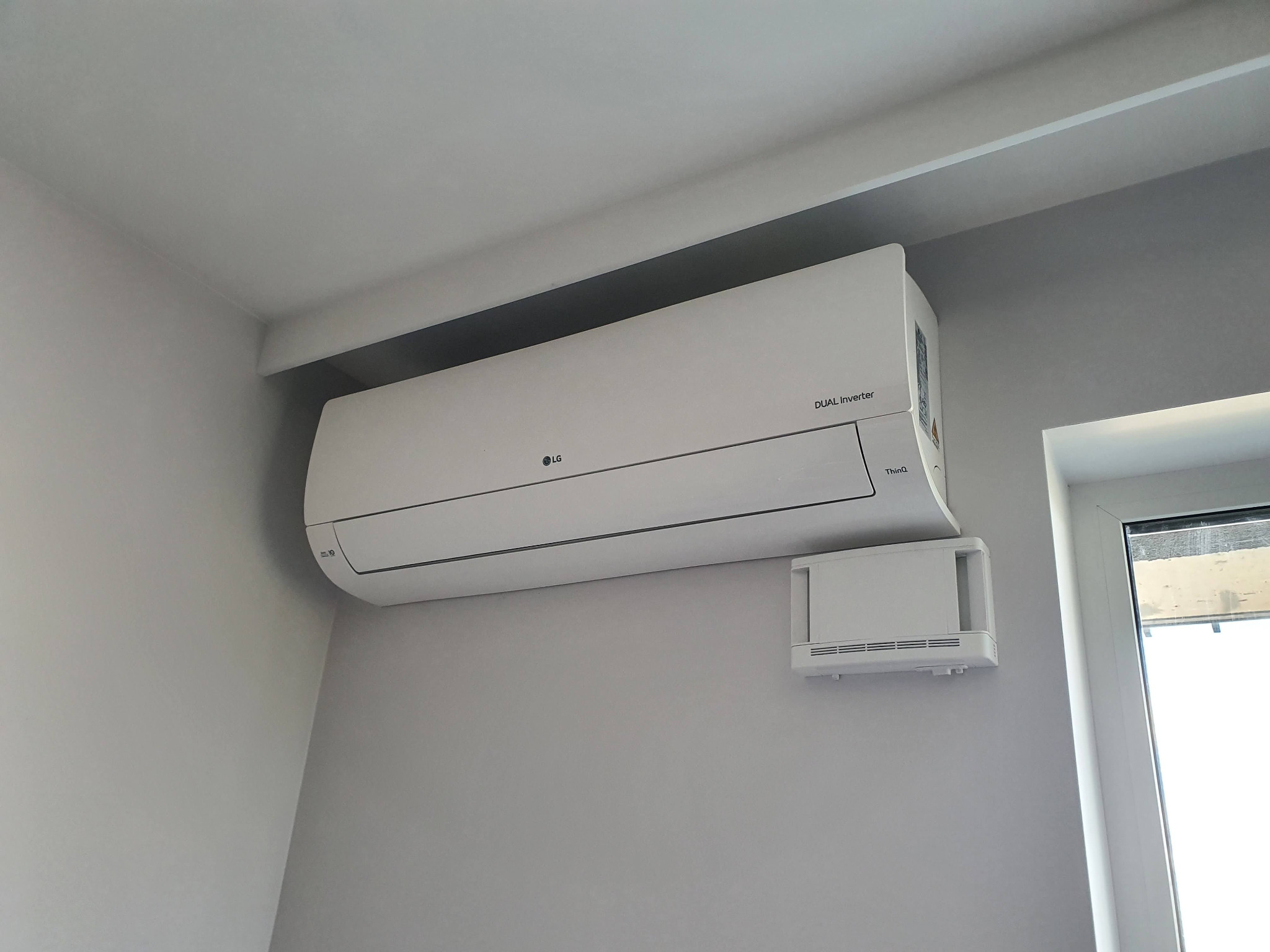 Biała jednostka wewnętrzna klimatyzacji LG Dual Inverter zamontowana na szarej ścianie w pomieszczeniu, widoczny również mały moduł sterujący i fragment okna.