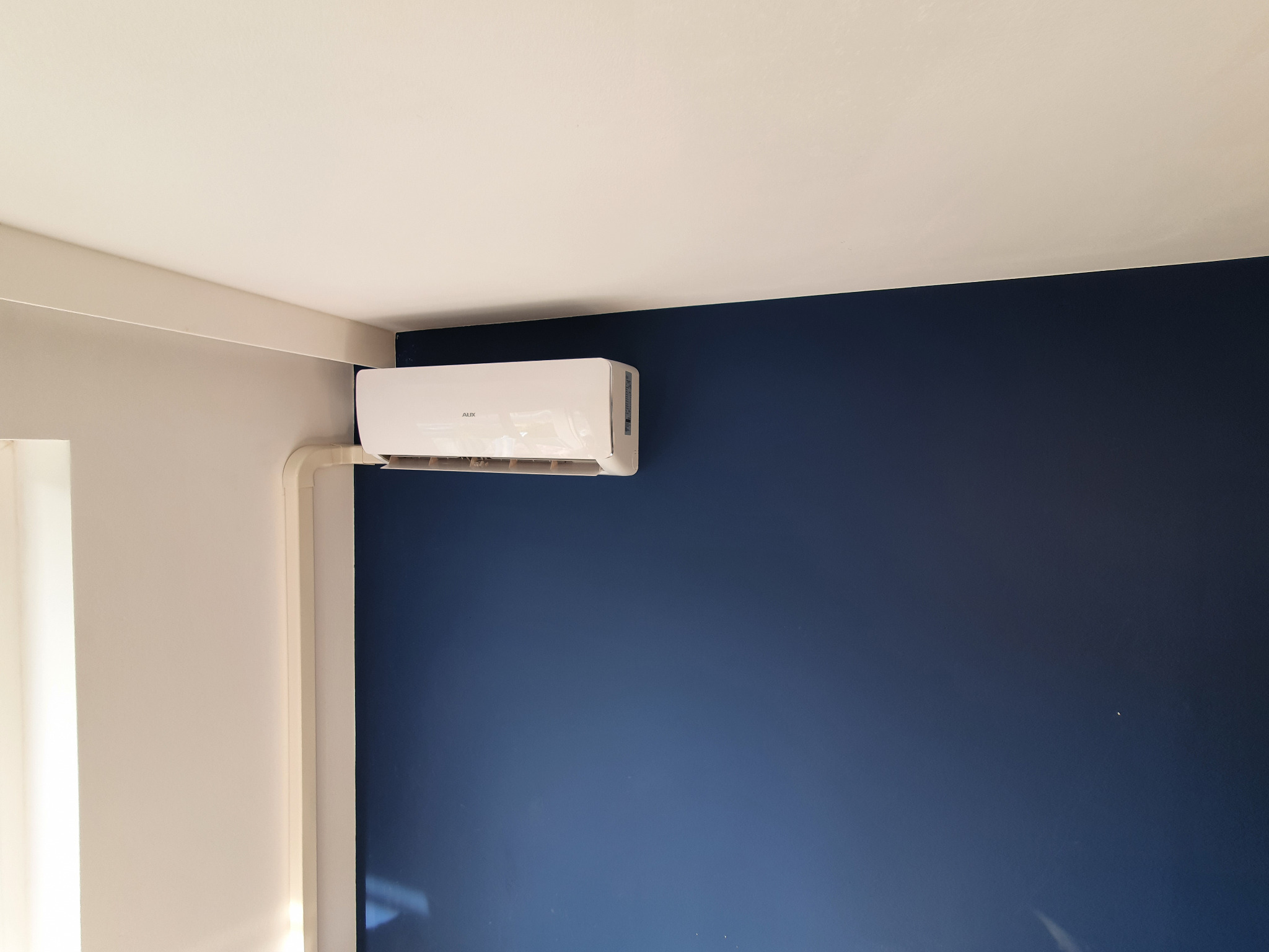 Biały klimatyzator AUX zamontowany na granatowej ścianie w rogu pokoju, z widocznymi białymi listwami przypodłogowymi i rurami instalacyjnymi.
