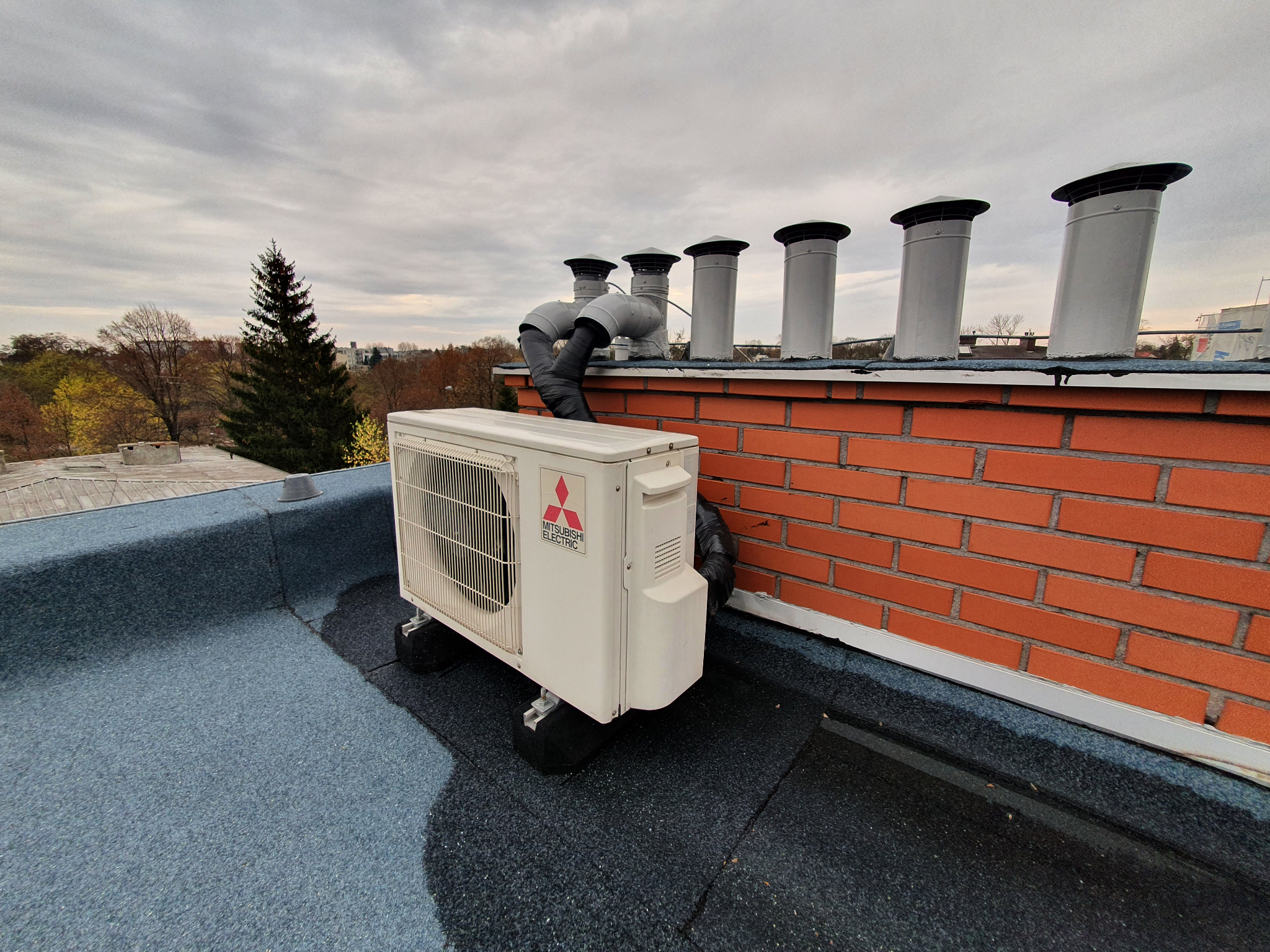 Biała jednostka zewnętrzna klimatyzacji Mitsubishi Electric zamontowana na płaskim dachu z papy, widoczne kominy wentylacyjne i ceglany mur, pochmurne niebo w tle.
