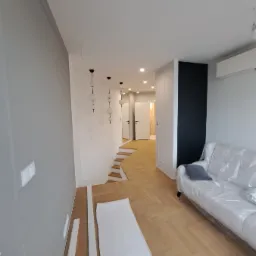 Nowoczesne wnętrze z parkietem w jodełkę, schodami i sofą zabezpieczoną folią, widoczne listwy przypodłogowe przed montażem.