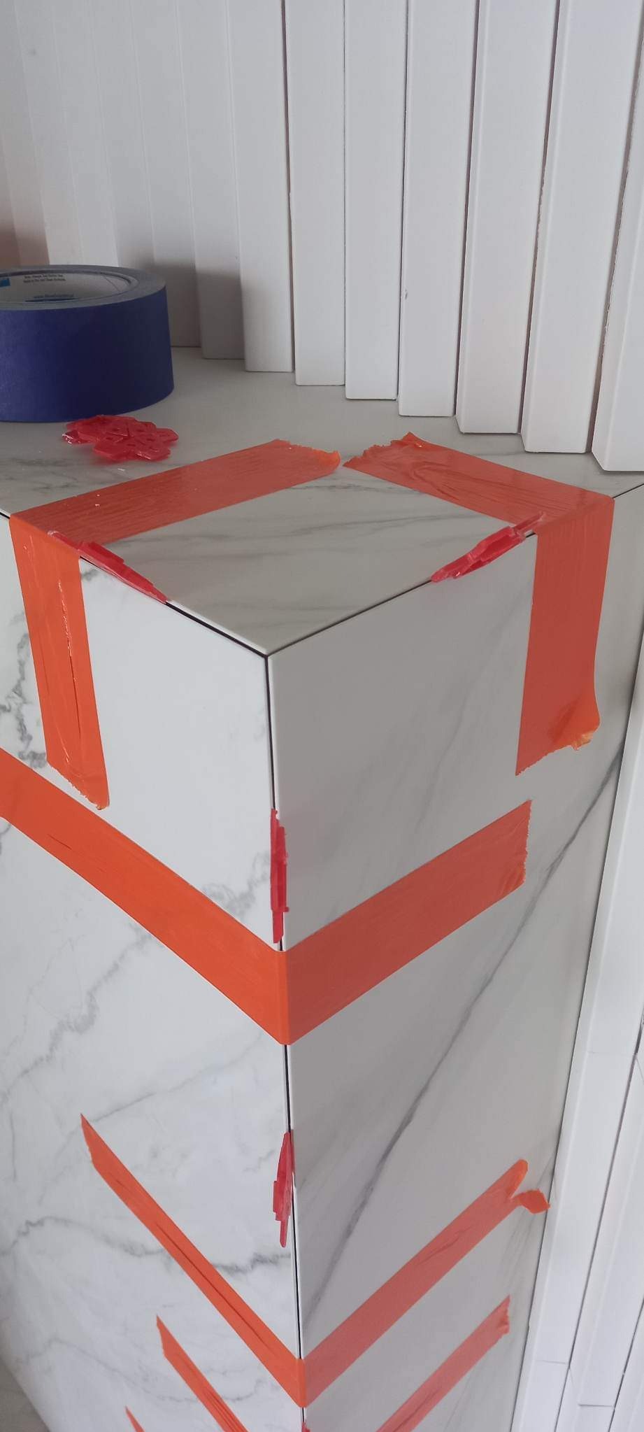 Fragment narożnika obłożonego białymi płytkami imitującymi marmur, zabezpieczonego pomarańczową taśmą malarską i czerwonymi krzyżykami dystansowymi; w tle widoczna rolka niebieskiej taśmy...