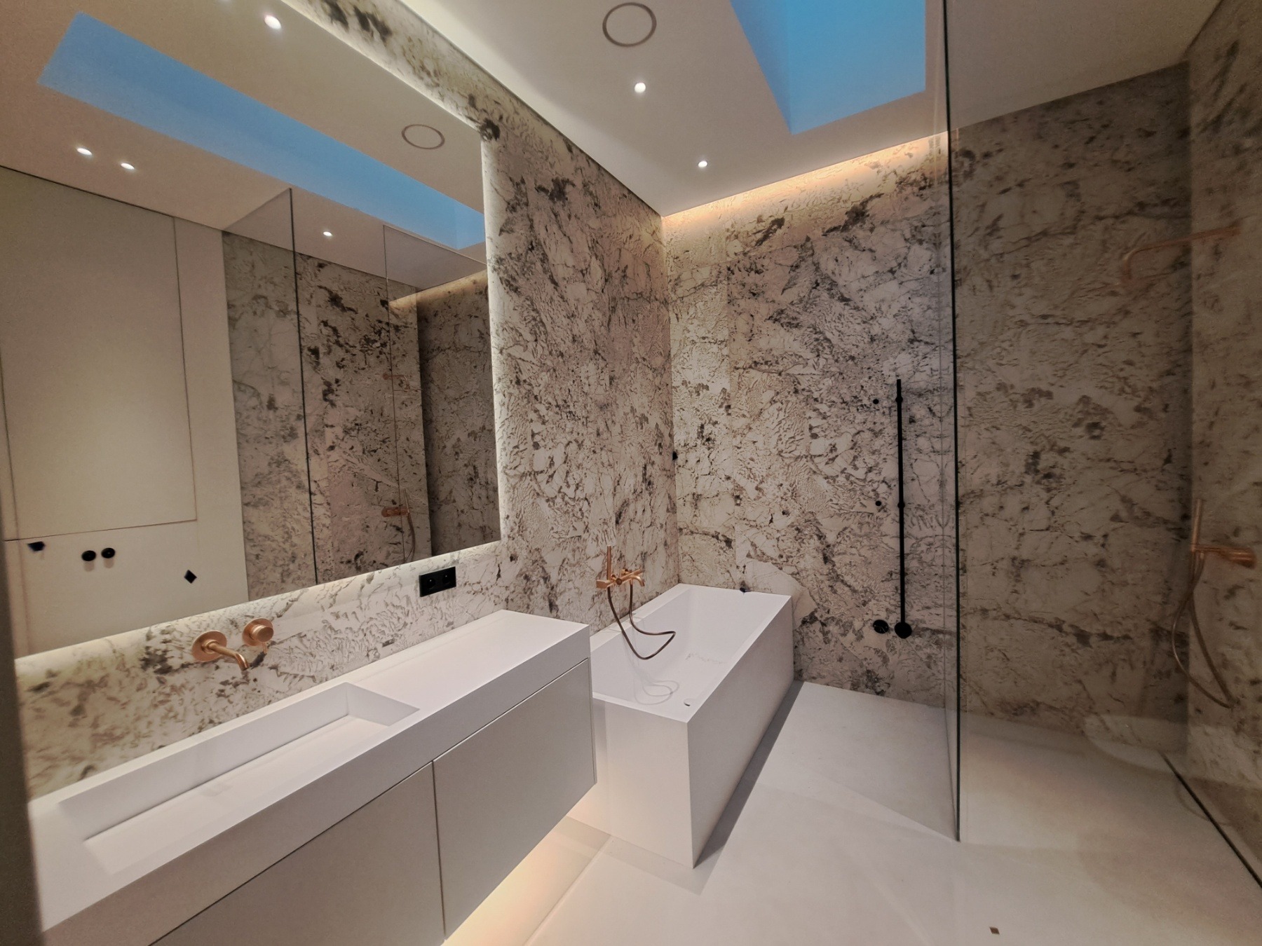 Nowoczesna łazienka z marmurowymi ścianami, białą armaturą i oświetleniem sufitowym imitującym naturalne światło dzienne.