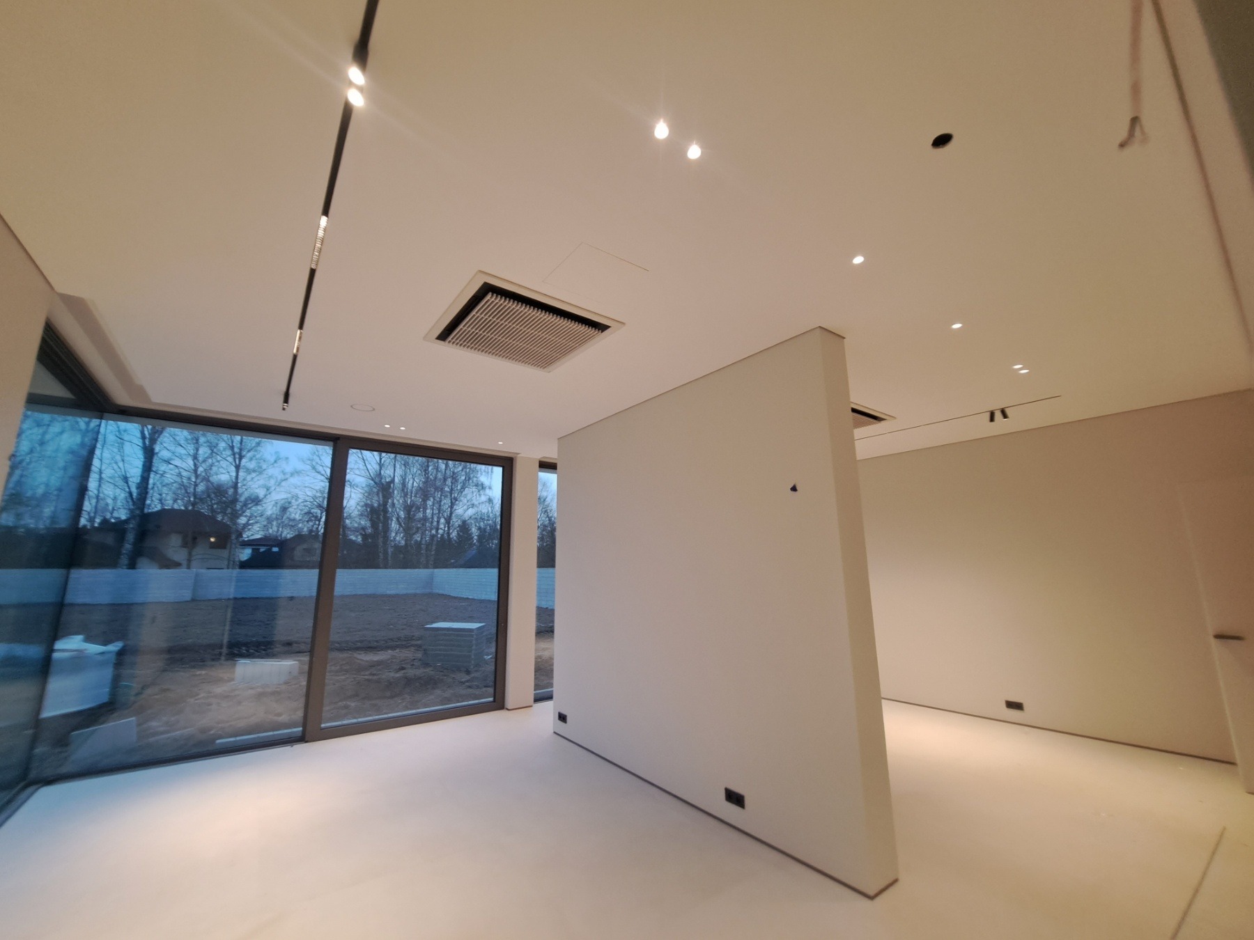 Nowoczesne wnętrze w trakcie wykańczania z dużym oknem, widok na zewnątrz, minimalistyczne oświetlenie sufitowe i ścianka działowa z widocznymi gniazdkami elektrycznymi.