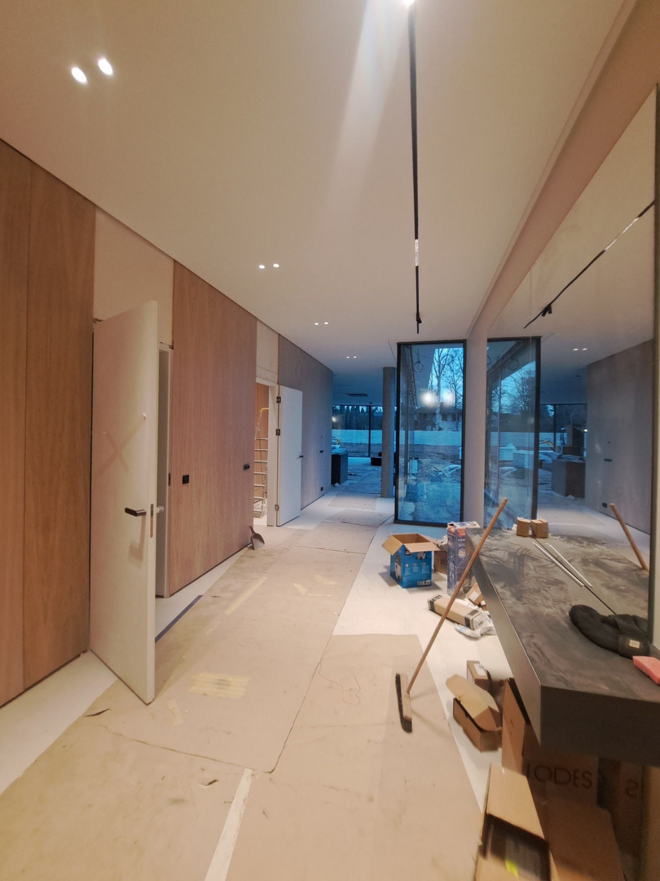 Wnętrze w trakcie prac wykończeniowych: zabezpieczona podłoga, zabudowa drewniana na ścianach, otwarte drzwi, widoczne narzędzia i materiały budowlane, duże okna z widokiem na zewnątrz.