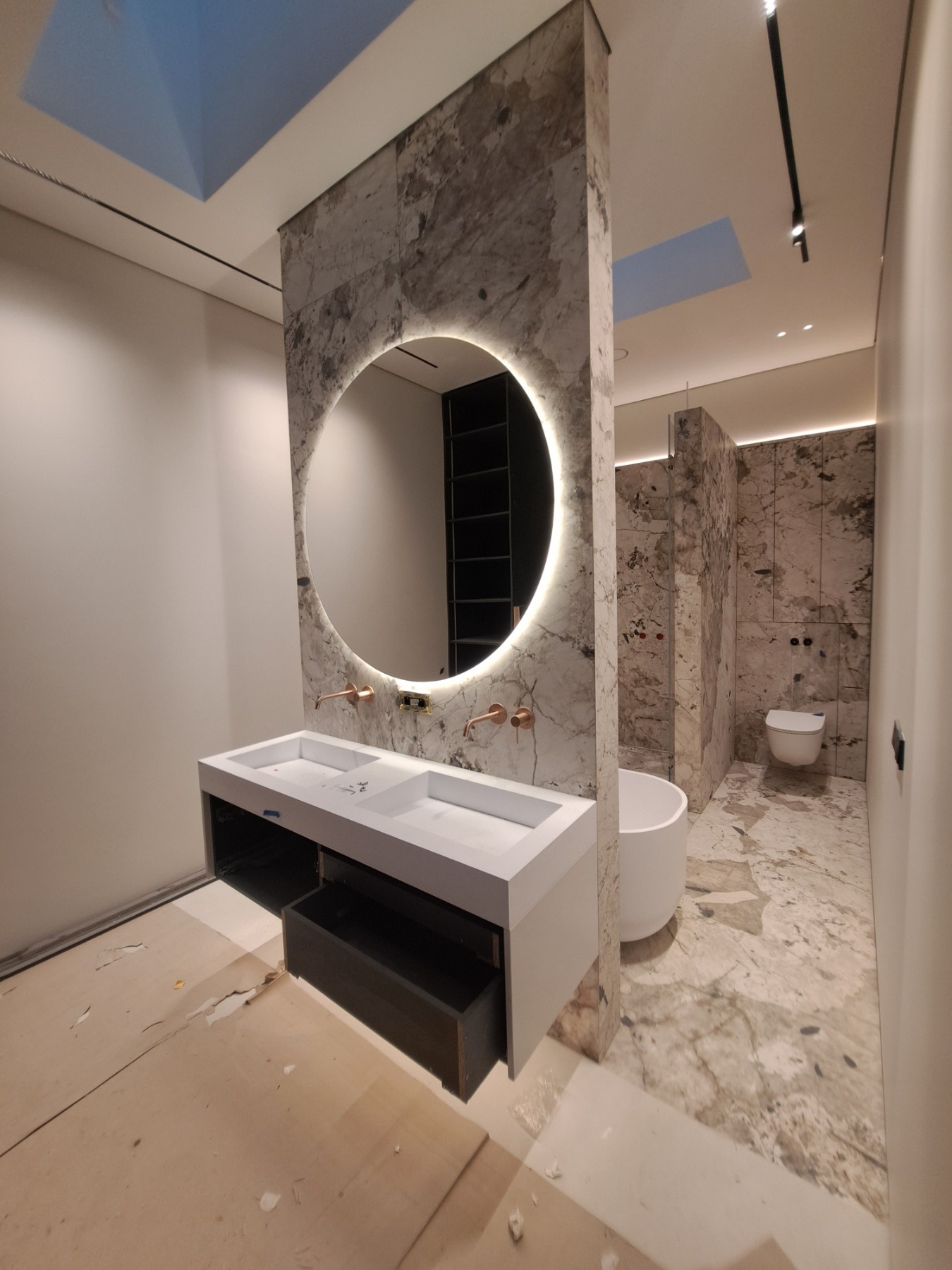 Nowoczesna łazienka w trakcie wykańczania: marmurowe ściany, okrągłe lustro z podświetleniem LED, biała umywalka z dwoma kranami w kolorze miedzi, wolnostojąca wanna i wisząca toaleta.