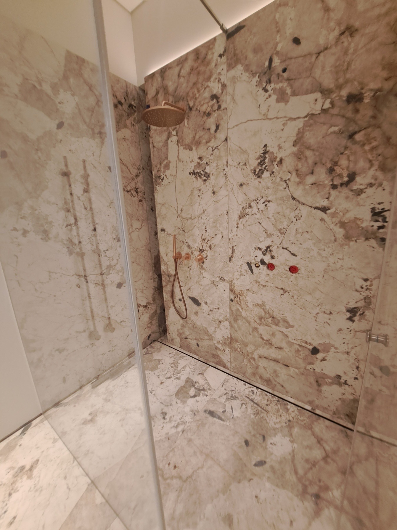 Kabina prysznicowa z marmurowymi płytami w odcieniach beżu i brązu, deszczownica i bateria w kolorze różowego złota, widoczne czerwone zaślepki na instalacji wodnej.