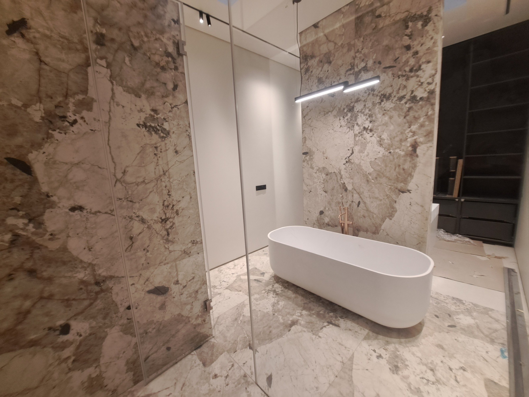 Nowoczesna łazienka w trakcie wykańczania z wolnostojącą białą wanną, marmurowymi płytami na ścianach i podłodze, minimalistyczną armaturą oraz oświetleniem LED, widoczne elementy konstrukcyjne...