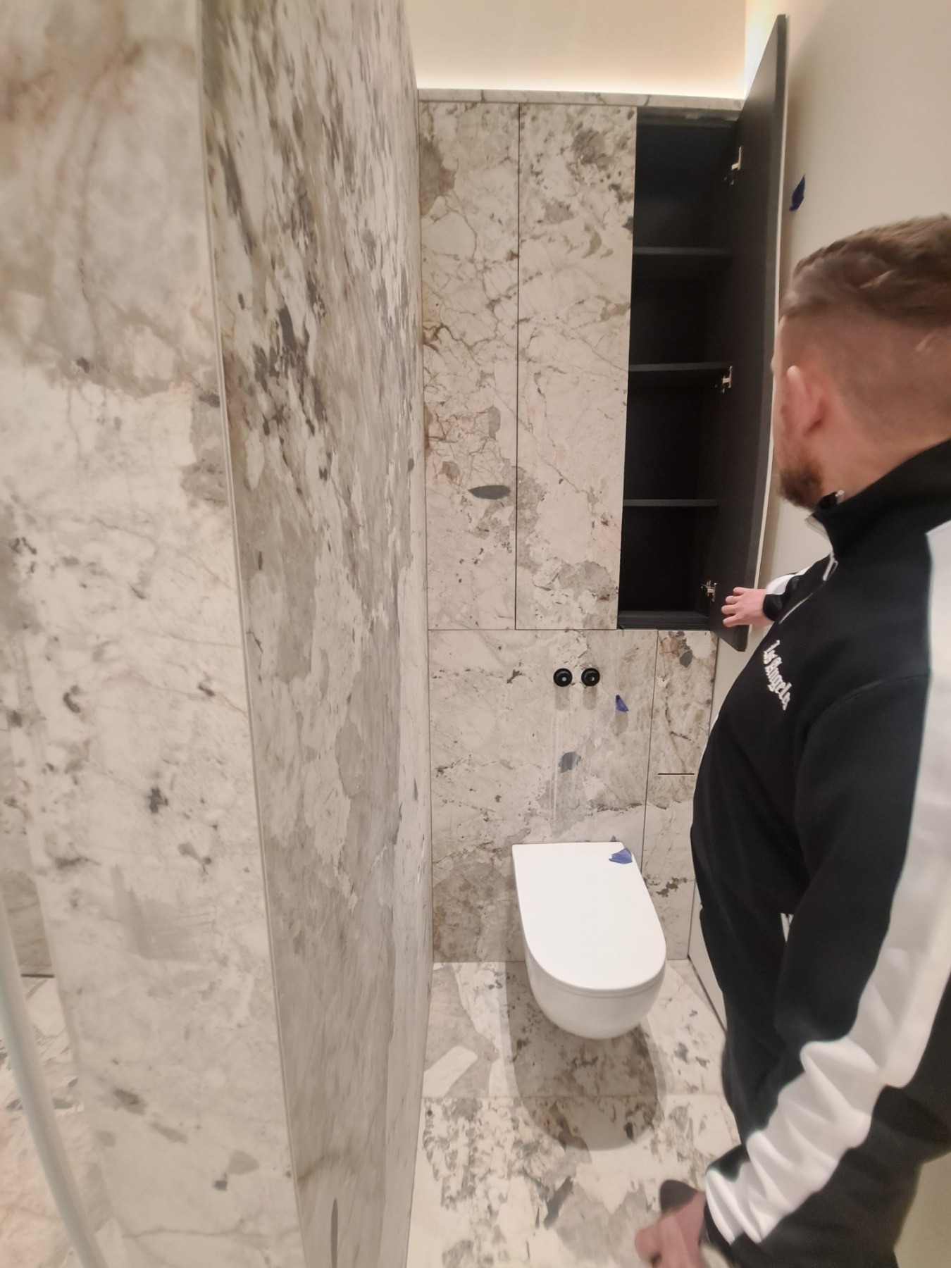 Wykończona łazienka z marmurowymi płytkami na ścianach i podłodze, wisząca toaleta z niebieskim zabezpieczeniem na desce, zabudowa szafkowa z otwartymi drzwiczkami, widoczny mężczyzna w bluzie z logo.