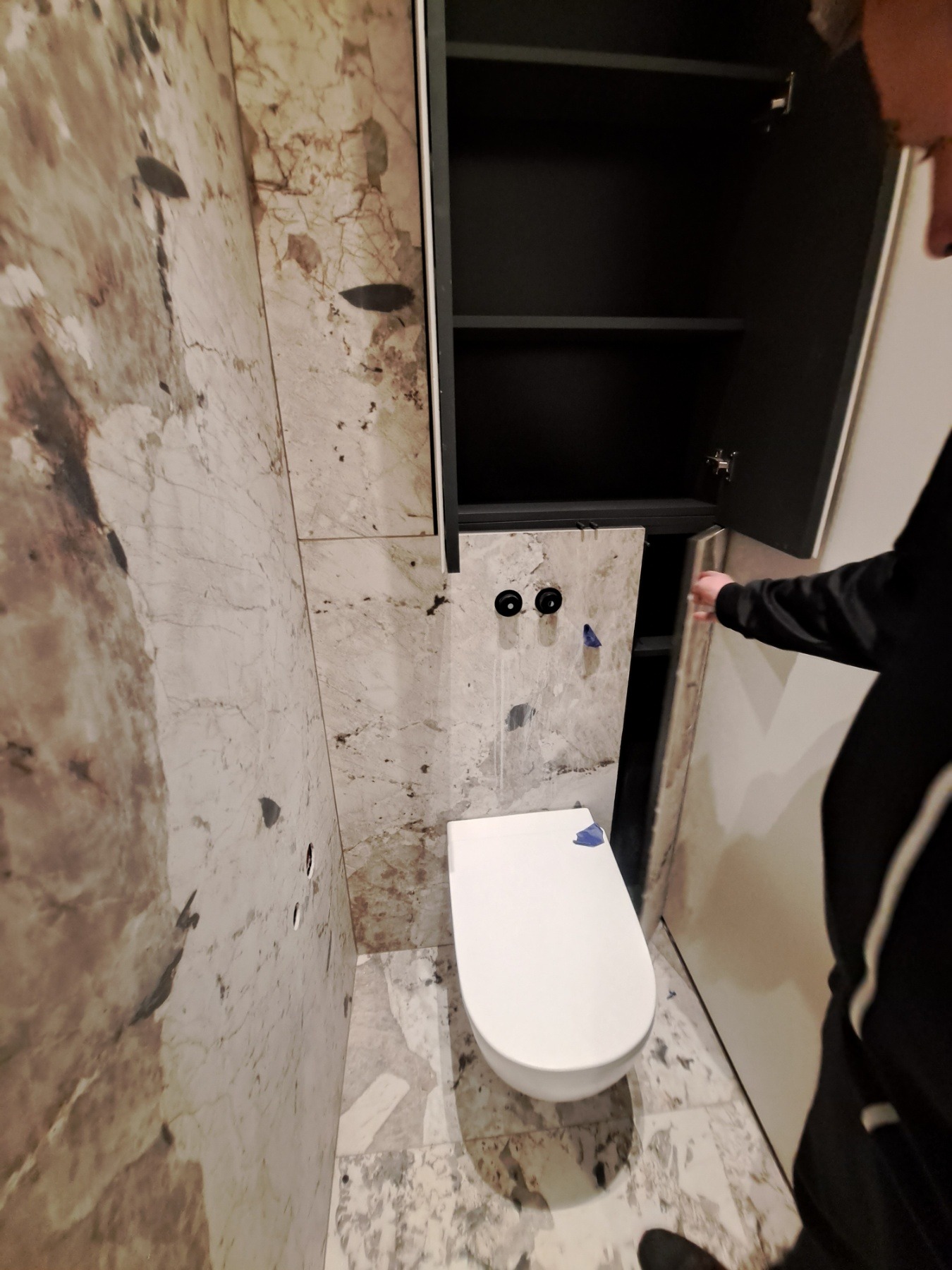 Wisząca toaleta z zabudową szafkową w łazience wykończonej marmurowymi płytami, widoczne przygotowane punkty instalacyjne i częściowo otwarte drzwiczki szafki.