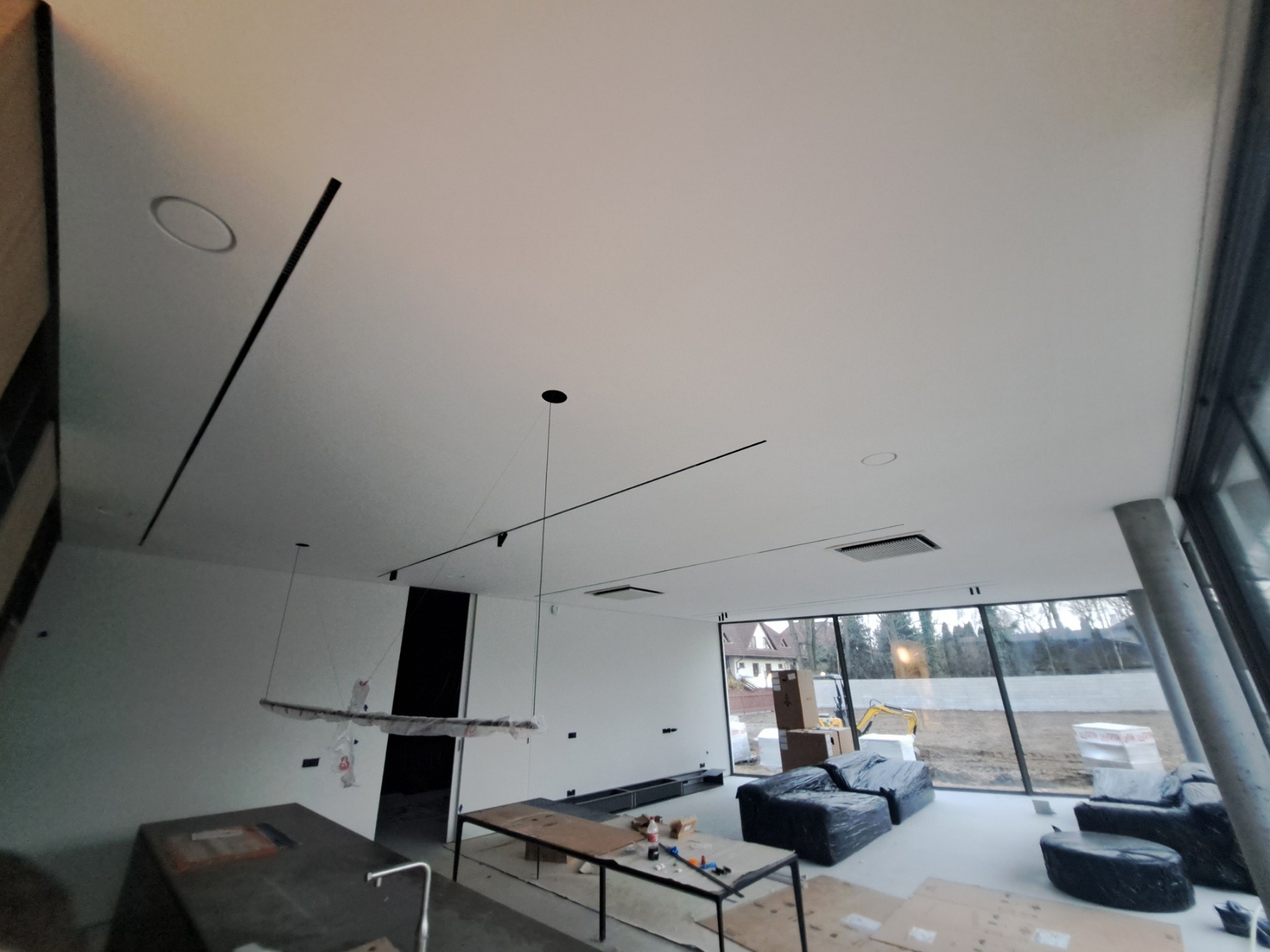 Wnętrze domu w trakcie prac wykończeniowych w Warszawie, widoczne minimalistyczne oświetlenie sufitowe, duże okna z widokiem na zewnątrz i meble zabezpieczone folią.