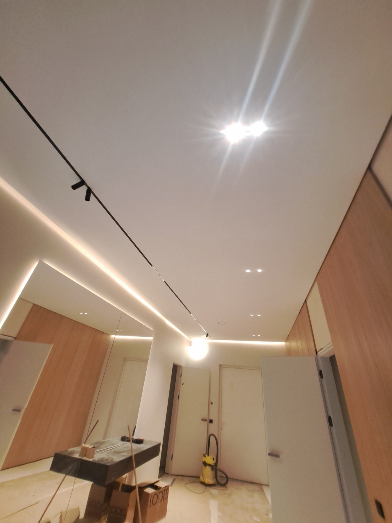 Nowoczesny korytarz w trakcie prac wykończeniowych, z oświetleniem LED na suficie, drewnianą okładziną ścian i widocznym odkurzaczem oraz materiałami budowlanymi na podłodze.