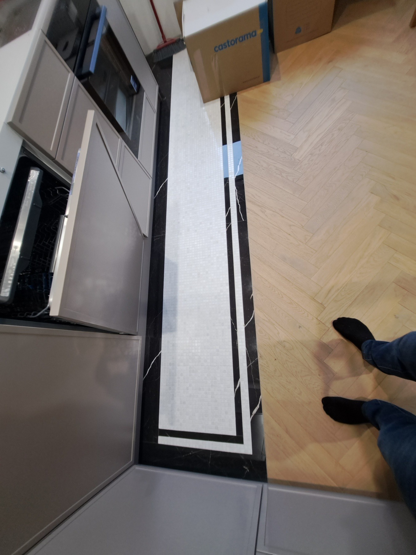 Kuchnia po remoncie: szare szafki z otwartymi drzwiczkami, podłoga z jasnego parkietu ułożonego w jodełkę, obok biała mozaika oddzielona czarnym pasem imitującym marmur, w tle karton Castorama.