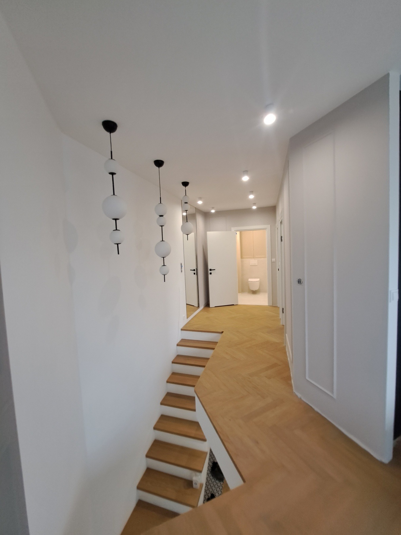 Nowoczesny korytarz z drewnianą podłogą ułożoną w jodełkę, białymi ścianami, schodami z drewnianymi stopniami, trzema wiszącymi lampami z kulistymi kloszami i widokiem na łazienkę z toaletą.