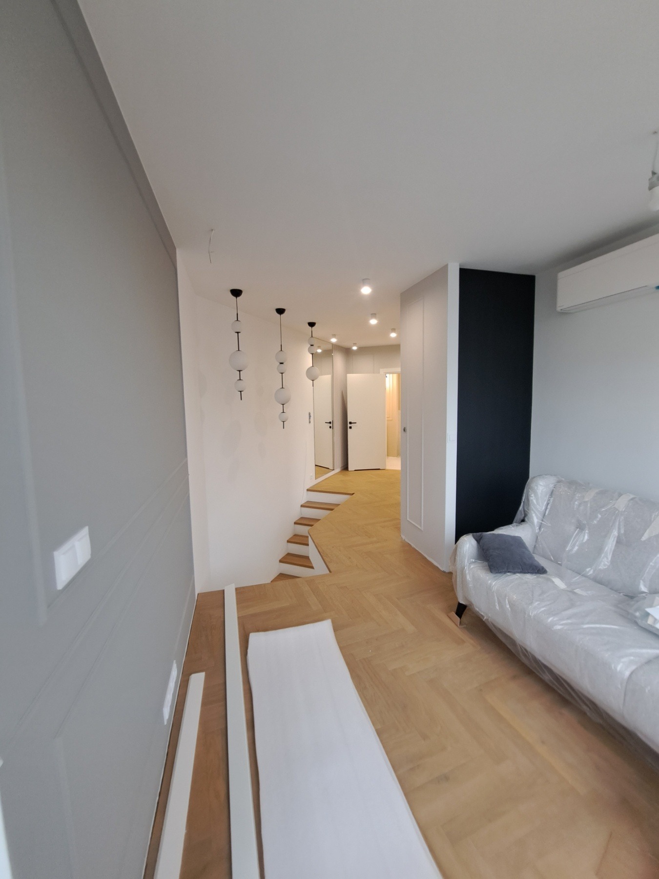 Nowoczesne wnętrze z parkietem w jodełkę, schodami i sofą zabezpieczoną folią, widoczne listwy przypodłogowe przed montażem.