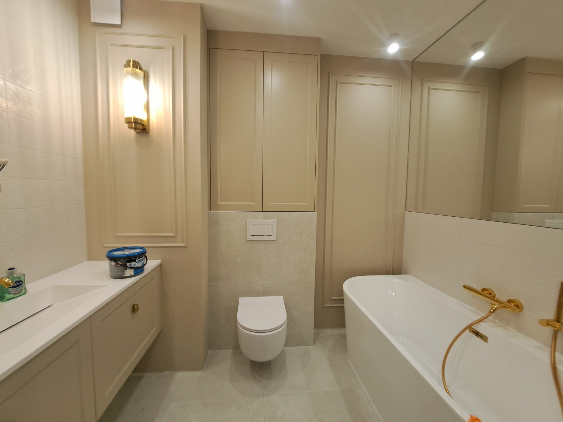 Elegancka łazienka w beżach z wanną, wiszącą toaletą i szafkami, uzupełniona oświetleniem w stylu glamour i złotą armaturą. Na blacie stoi wiaderko z farbą.