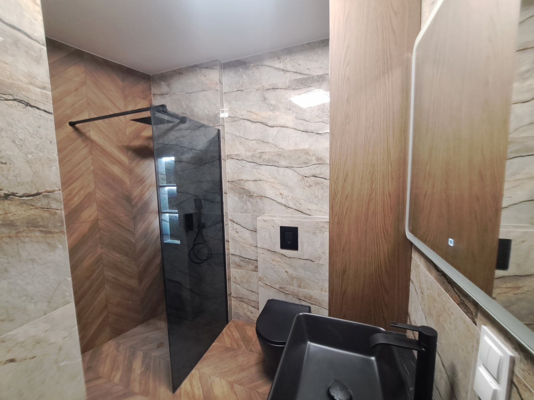 Nowoczesna łazienka z kabiną prysznicową typu walk-in z czarnymi detalami, umywalką nablatową i toaletą w kolorze czarnym, wykończona płytkami imitującymi marmur i drewno.