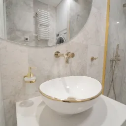 Elegancka łazienka z umywalką nablatową w białej misie z złotym rantem, złota armatura, marmurowe płytki ścienne i okrągłe lustro, w tle pralka.