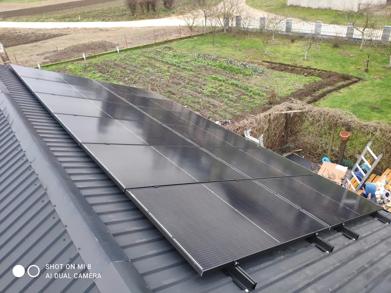 Panele fotowoltaiczne na metalowym dachu z widocznym ogrodem w tle, ujęcie z góry, widoczne elementy montażowe paneli.