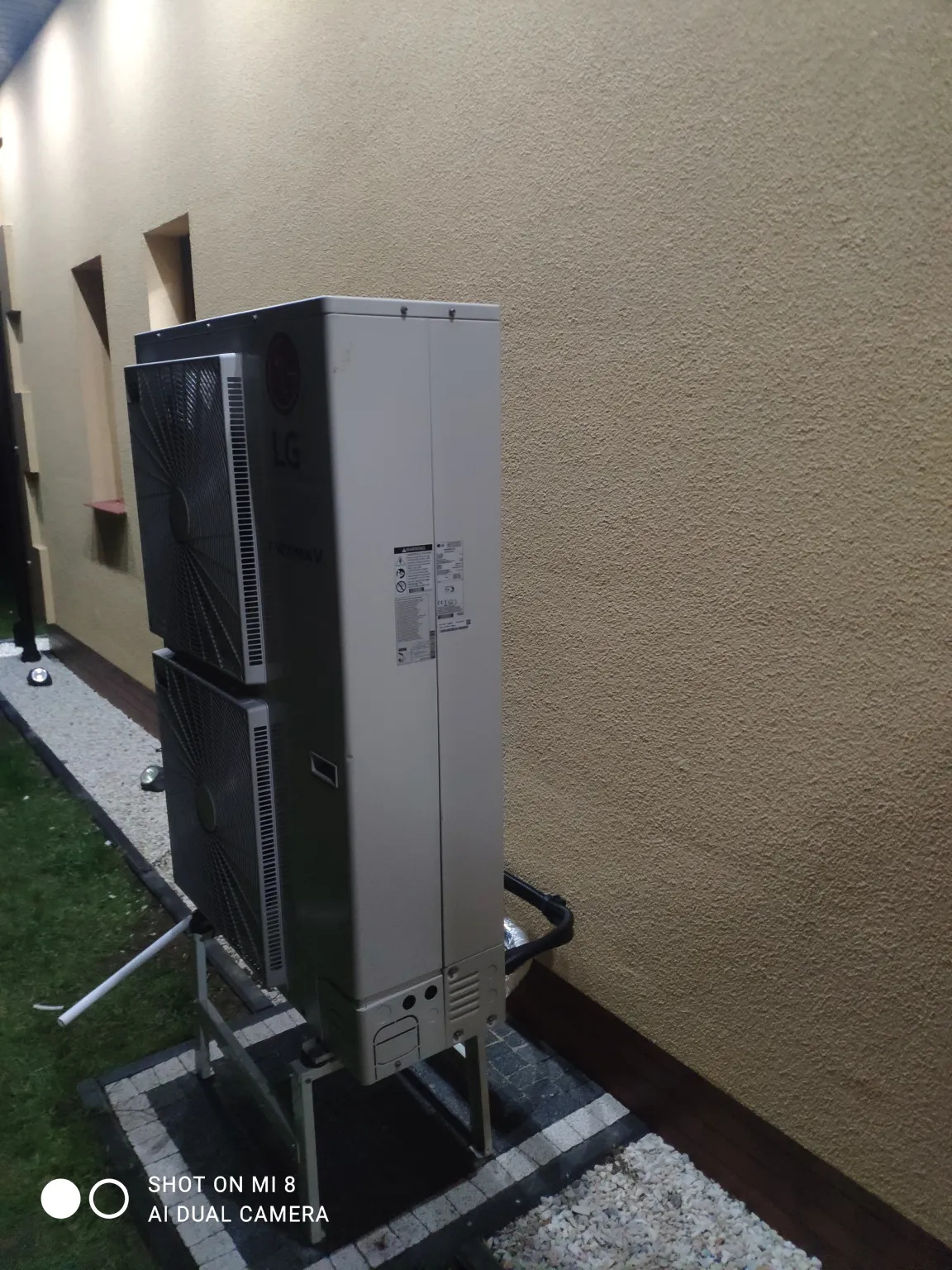 Zewnętrzna jednostka pompy ciepła LG Therma V na metalowym stelażu, zainstalowana przy beżowej ścianie budynku z małymi okienkami i kamiennym obrzeżem, widok z boku.