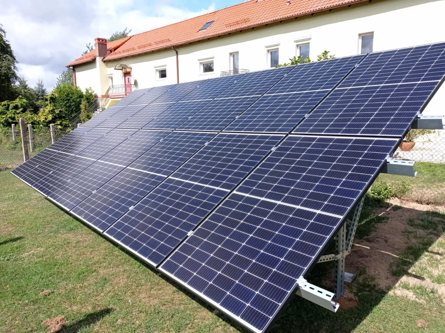 Instalacja paneli fotowoltaicznych na trawniku przed domem z czerwoną dachówką, widoczne metalowe konstrukcje wsporcze i ogrodzenie z siatki.