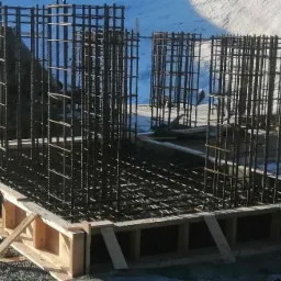 Zbrojenie fundamentu z drewnianym szalunkiem w zimowej scenerii, widoczne pionowe i poziome pręty zbrojeniowe, śnieg w tle.