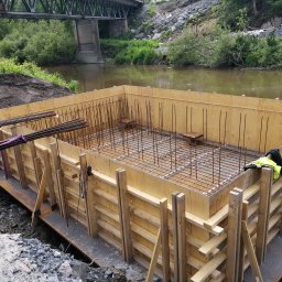 Grzegorz Kowalczyk GLOBAL-BUD - Szczegółowe ujęcie szalunku zbrojeniowego na fundamencie, z widoczną konstrukcją stalową i drewnianą formą, na tle rzeki i mostu. Prace ziemne w toku.