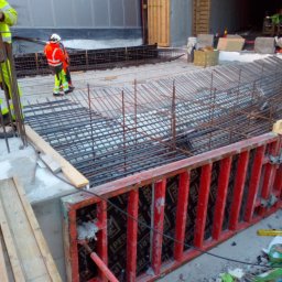 Grzegorz Kowalczyk GLOBAL-BUD - Zbrojenie fundamentów w czerwonym szalunku, widoczni pracownicy w odblaskowych ubraniach. Konstrukcja z metalowych prętów na budowie.