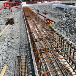 Grzegorz Kowalczyk GLOBAL-BUD - Zbrojenie fundamentów w wykopie, widok z góry. Stalowe pręty tworzą szkielet konstrukcji, przygotowany do zalania betonem. W tle gruz i elementy infrastruktury.