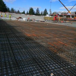 Grzegorz Kowalczyk GLOBAL-BUD - Szeroki kadr na rozległą siatkę zbrojeniową na budowie, z dźwigiem w tle i pracownikami w odblaskowych ubraniach. Niebo błękitne z chmurami.