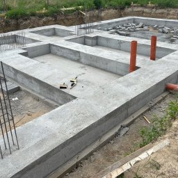 Grzegorz Kowalczyk GLOBAL-BUD - Zalane betonem fundamenty z wystającymi prętami zbrojeniowymi i pomarańczowymi rurami kanalizacyjnymi. Na fundamencie leżą bloczki betonowe.