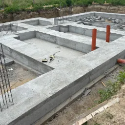 Zalane betonem fundamenty z wystającymi prętami zbrojeniowymi i pomarańczowymi rurami kanalizacyjnymi. Na fundamencie leżą bloczki betonowe.