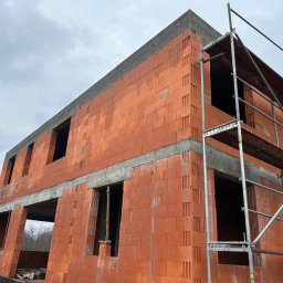 Grzegorz Kowalczyk GLOBAL-BUD - Surowy stan dwukondygnacyjnego domu z czerwonej cegły ceramicznej, z widocznymi otworami okiennymi i betonowymi nadprożami, otoczonego rusztowaniem.